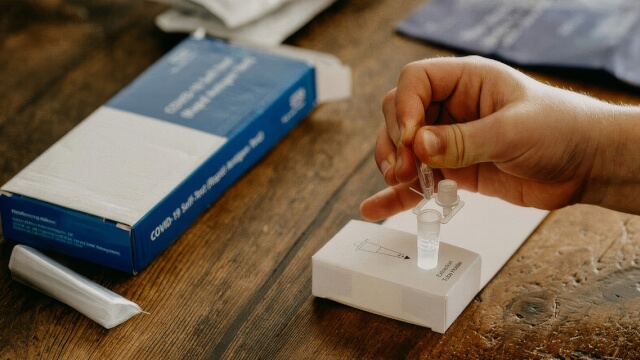 A person taking a COVID-19 rapid antigen test, with the test box on a wooden table